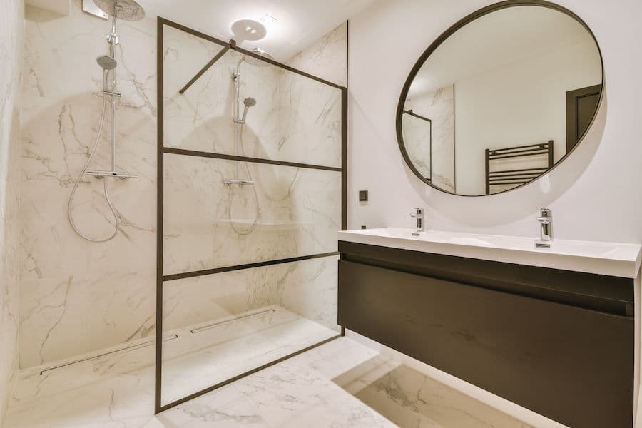 Modern bathroom with marble-tiled shower enclosure, black-framed glass doors, dark vanity with white countertop, round mirror, and heated towel rack