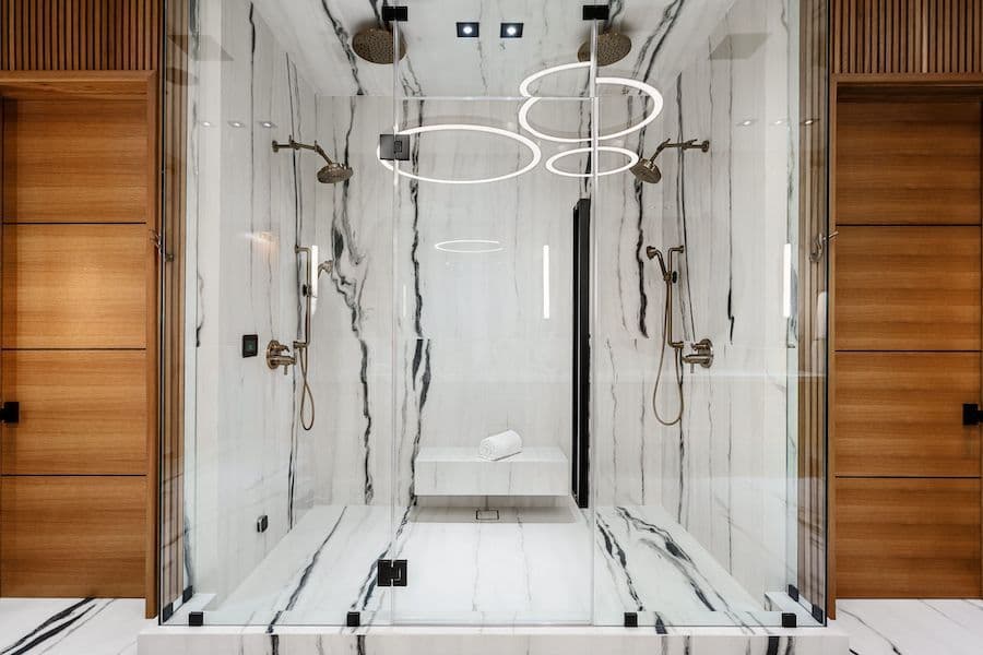 Modern luxury walk-in shower with marble-pattern tile walls, dual showerheads, neon ring lighting fixture, and frameless glass enclosure with black hardware
