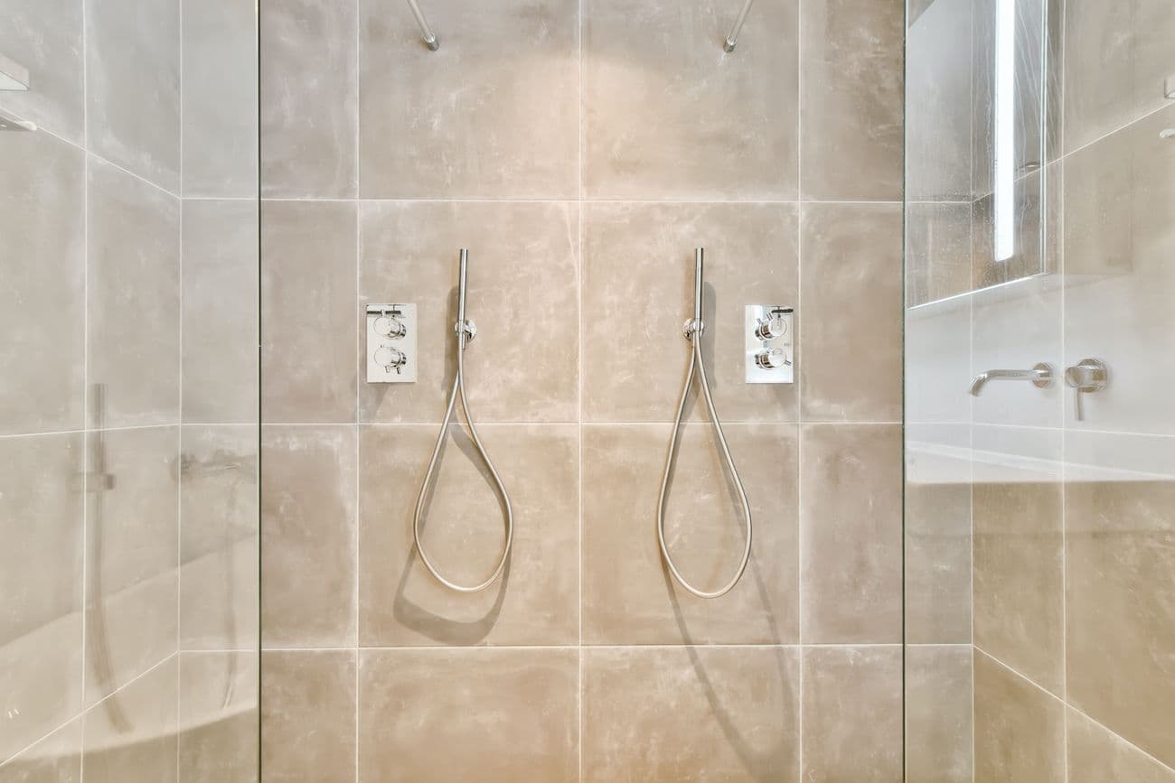 Luxury bathroom with dual rainfall showerheads and chrome fixtures in a walk-in shower with beige marble-look tile walls and glass enclosure