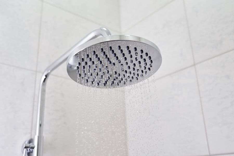 Round silver showerhead with multiple spray holes mounted on adjustable arm, dispensing water in bathroom with white tile walls