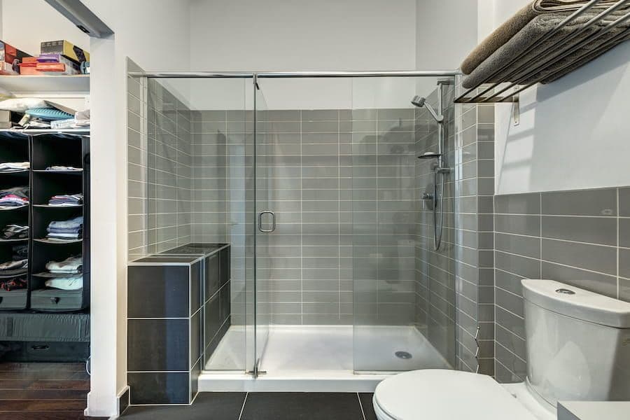 Modern bathroom with walk-in shower enclosed by glass door, gray tile walls, white toilet, and built-in storage shelving with folded linens on left